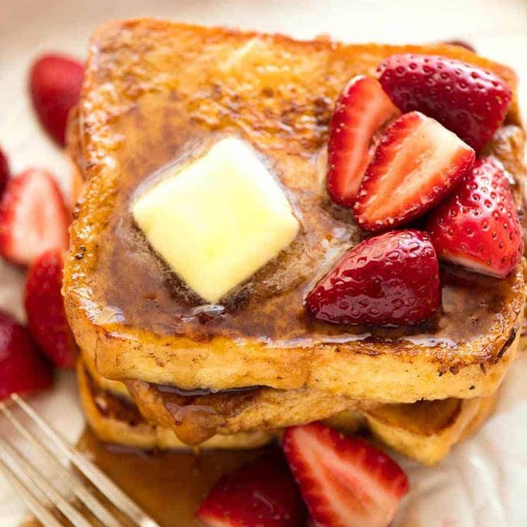 Pile of French Toast on a rustic white plate, doused with maple syrup and topped with a pat of melting butter and strawberries.