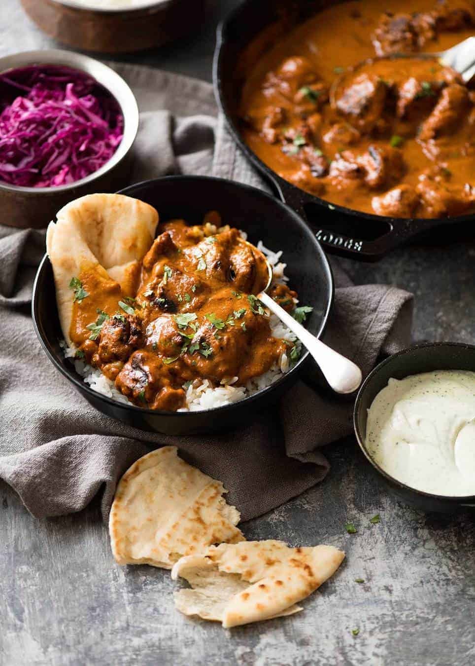Photo of chicken tikka masala on basmati rice in a dark rustic bowl with a piece of naan wedged in on the side. Pickled red cabbage and skillet of Chicken Tikka Masala in the background.